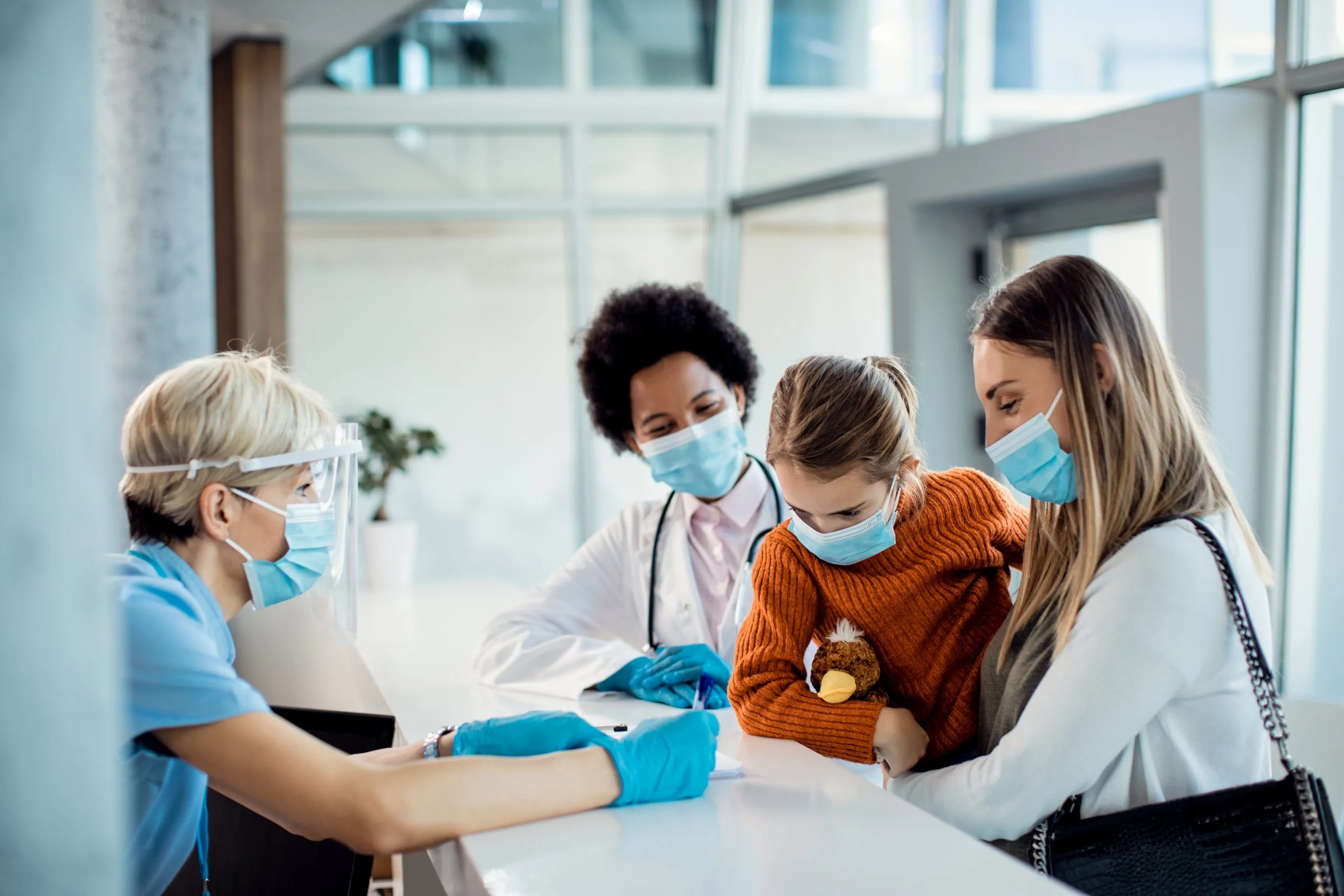 Little girl and her mother talking to a nurse at reception desk in the hospital during COVID-19 pandemic.