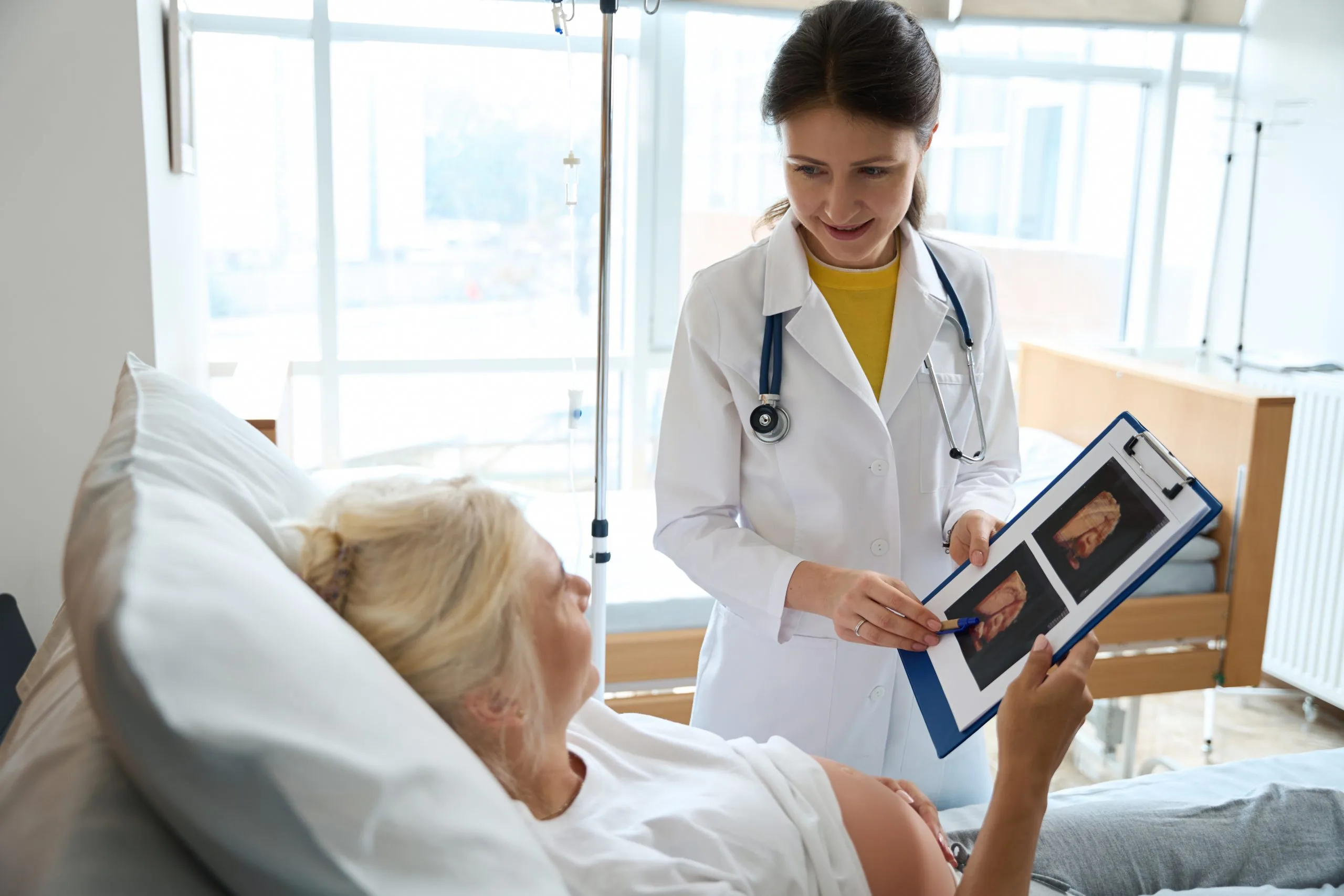 European female nurse with ultrasound photo and adult caucasian pregnant woman on medical bed looking at each other in maternity hospital